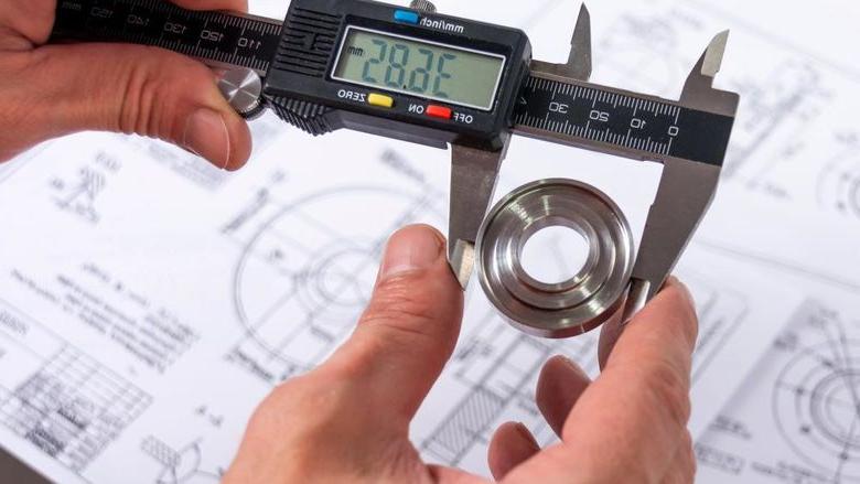 A quality engineer measuring a production part with calipers to ensure its compliance, with a reference diagram in the background.